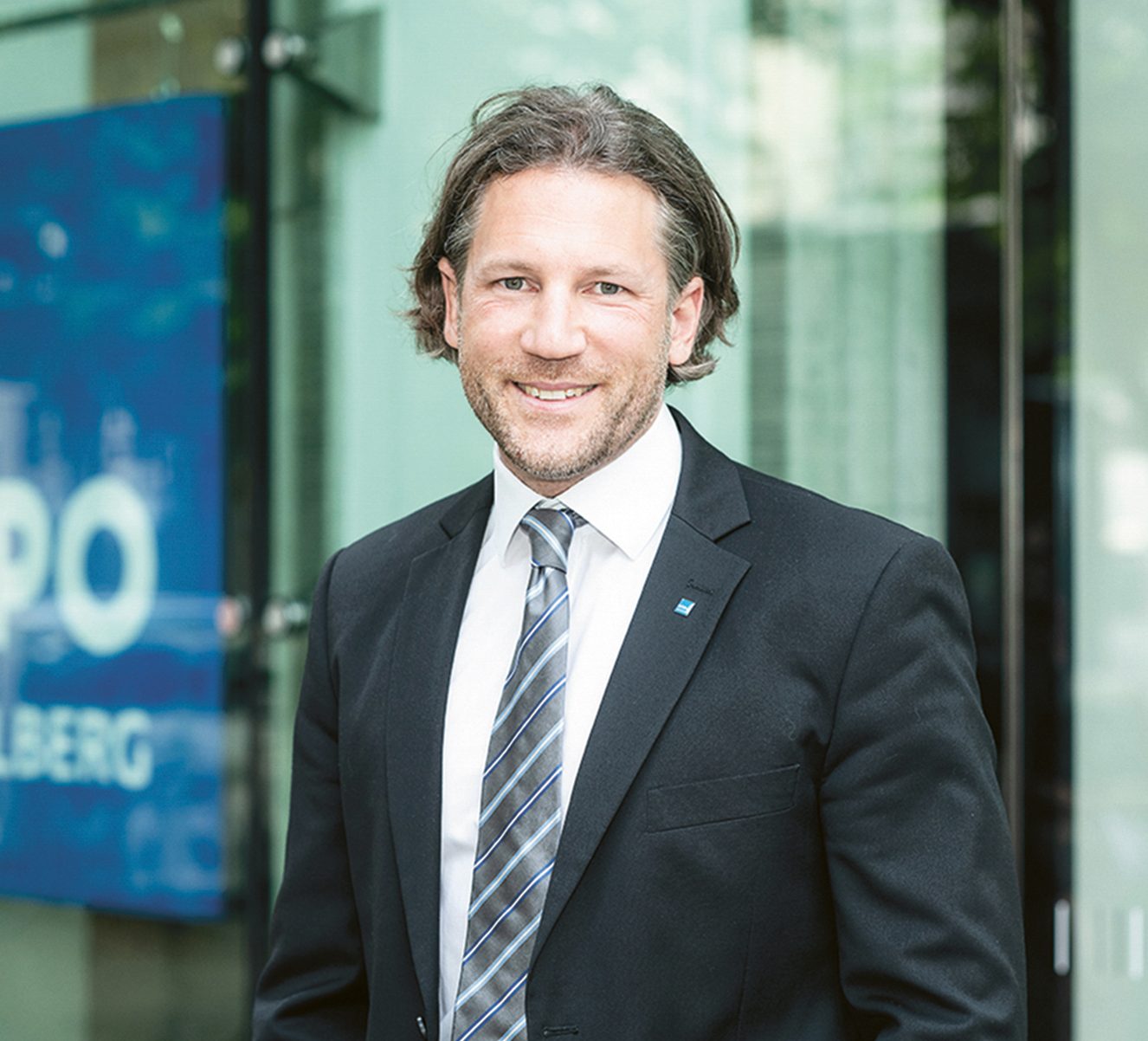 Ein lächelnder Mann in Anzug und Krawatte, der vor einem Bürogebäude der Hypo Vorarlberg steht.