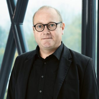Ein Mann mittleren Alters mit Brille, der ein schwarzes Hemd und einen Blazer trägt.