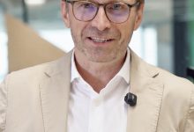 Ein Mann in einem hellbraunen Blazer und mit Brille spricht in die Kamera; er trägt ein kleines Ansteckmikrofon.