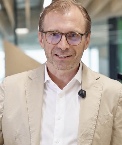 Ein Mann in einem hellbraunen Blazer und mit Brille spricht in die Kamera; er trägt ein kleines Ansteckmikrofon.