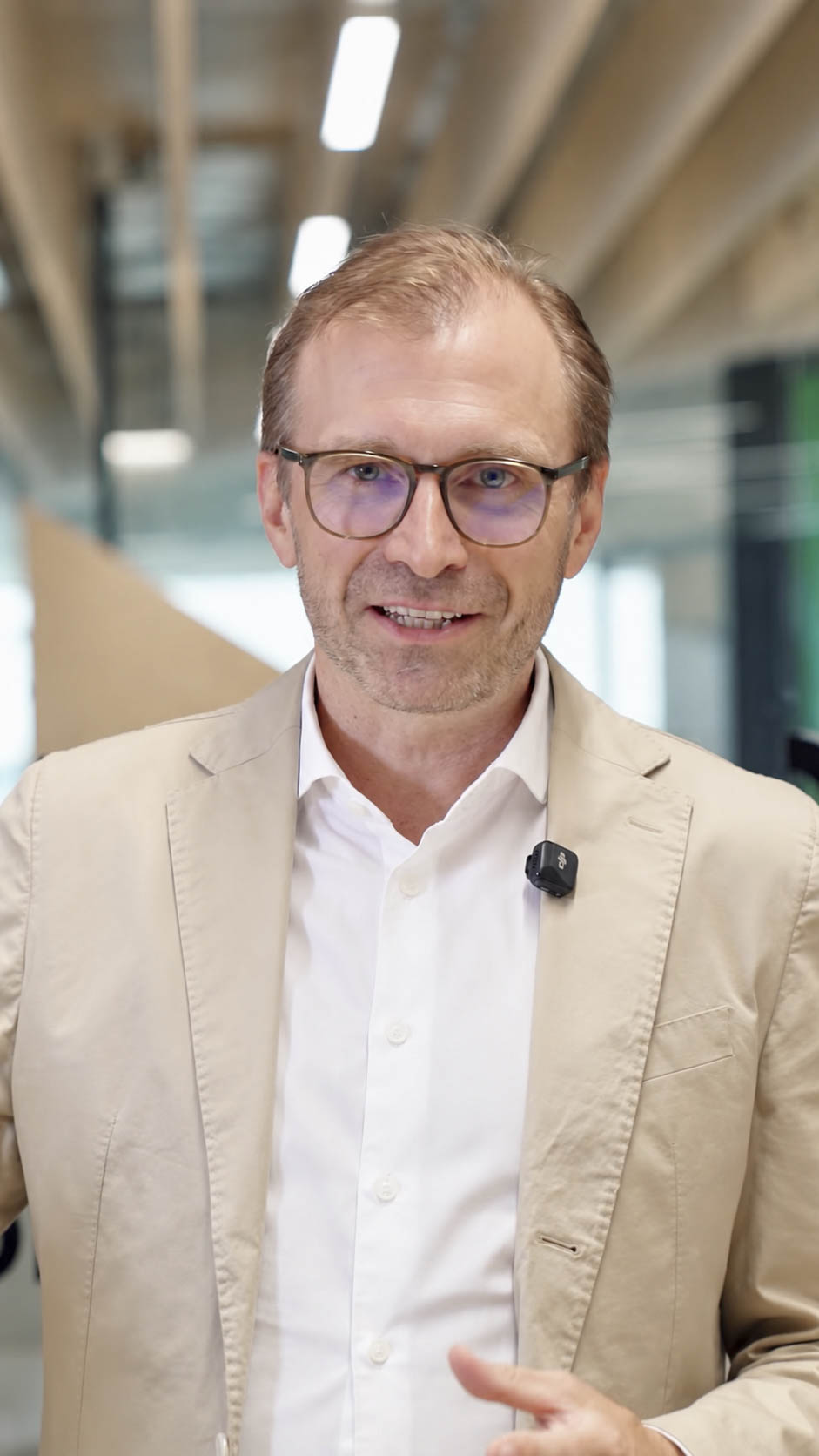 Ein Mann in einem hellbraunen Blazer und mit Brille spricht in die Kamera; er trägt ein kleines Ansteckmikrofon.