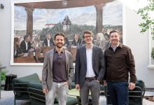 Drei Männer lächeln vor einem Wandbild, das Menschen an einem Tisch zeigt, die „Österreichs bester Nachwuchs-Hochbauer 2026“ feiern.