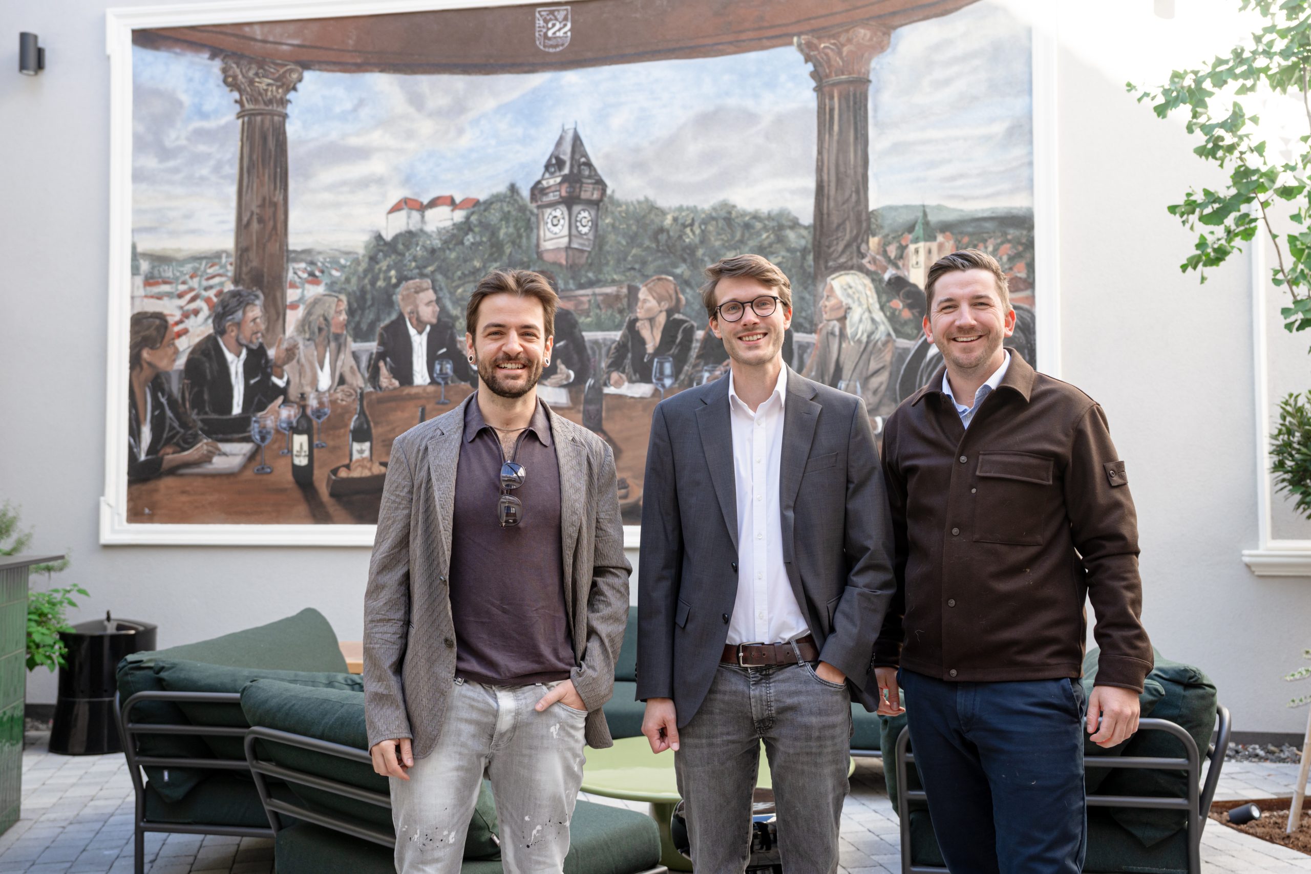 Drei Männer lächeln vor einem Wandbild, das Menschen an einem Tisch zeigt, die „Österreichs bester Nachwuchs-Hochbauer 2026“ feiern.