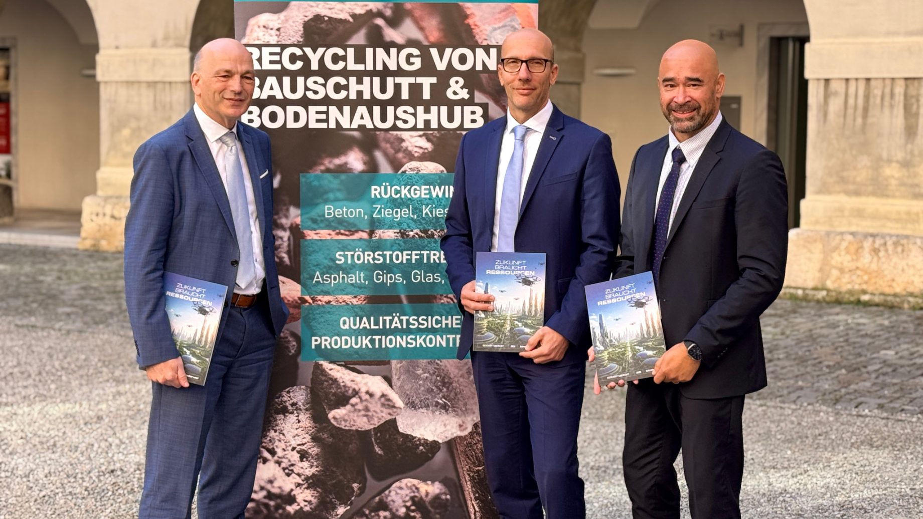 Drei Männer in Anzügen stehen vor einem Banner von Binder+Co Forschung und Entwicklung zum Thema Bauabfallrecycling und halten jeweils eine Broschüre in der Hand.