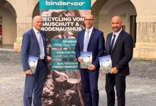 Drei Männer in Anzügen stehen vor einem Banner von Binder+Co Forschung und Entwicklung zum Thema Bauabfallrecycling und halten jeweils eine Broschüre in der Hand.