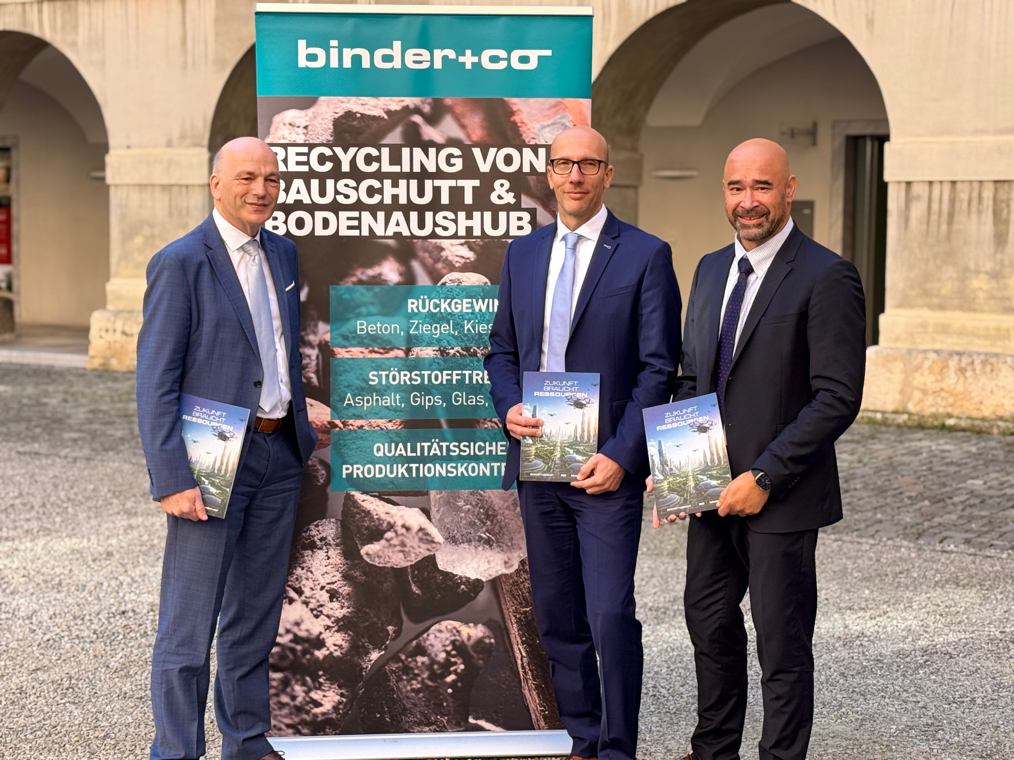 Drei Männer in Anzügen stehen vor einem Banner von Binder+Co Forschung und Entwicklung zum Thema Bauabfallrecycling und halten jeweils eine Broschüre in der Hand.