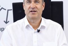 Ein Mann mittleren Alters in einem weißen Hemd sitzt an einem Schreibtisch vor einer SFG-Markenhintergrundkulisse.