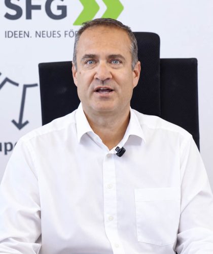 Ein Mann mittleren Alters in einem weißen Hemd sitzt an einem Schreibtisch vor einer SFG-Markenhintergrundkulisse.