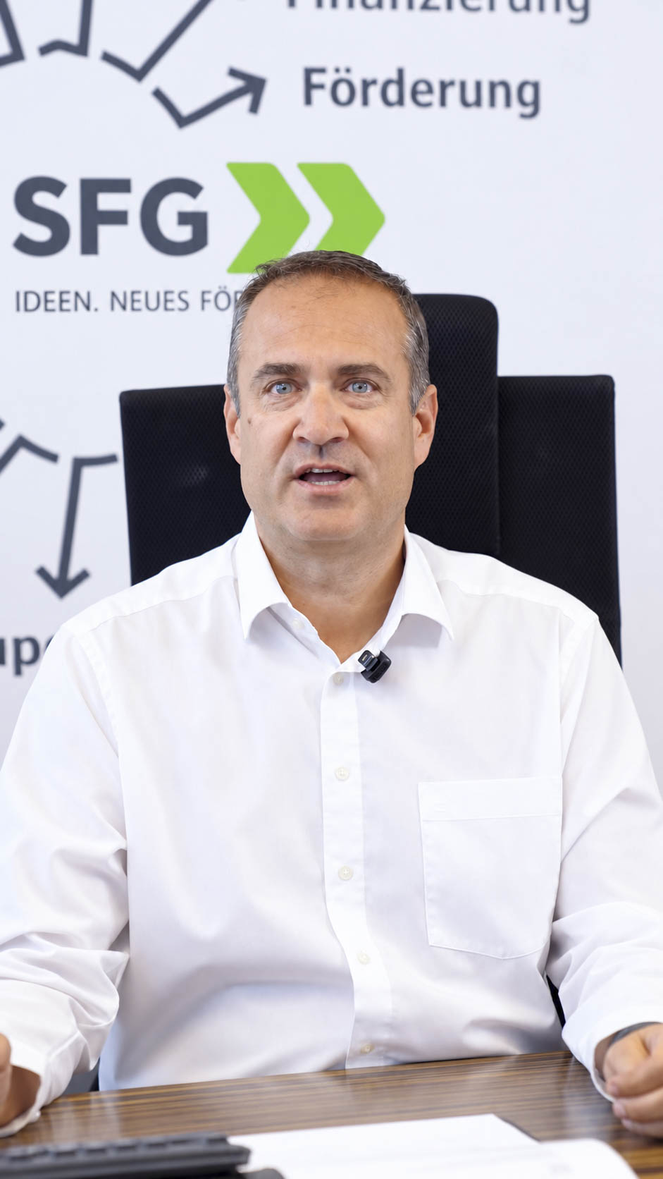 Ein Mann mittleren Alters in einem weißen Hemd sitzt an einem Schreibtisch vor einer SFG-Markenhintergrundkulisse.