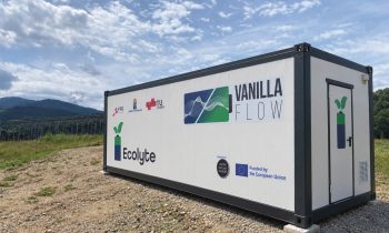 Ein mit „Vanilla Flow“ und „Ecolyte“ beschrifteter Versandcontainer steht unter blauem Himmel auf einem Schotterfeld.