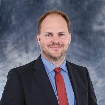 Ein Mann, der einen dunklen Anzug, ein blaues Hemd mit Knöpfen und eine rote Krawatte trägt.