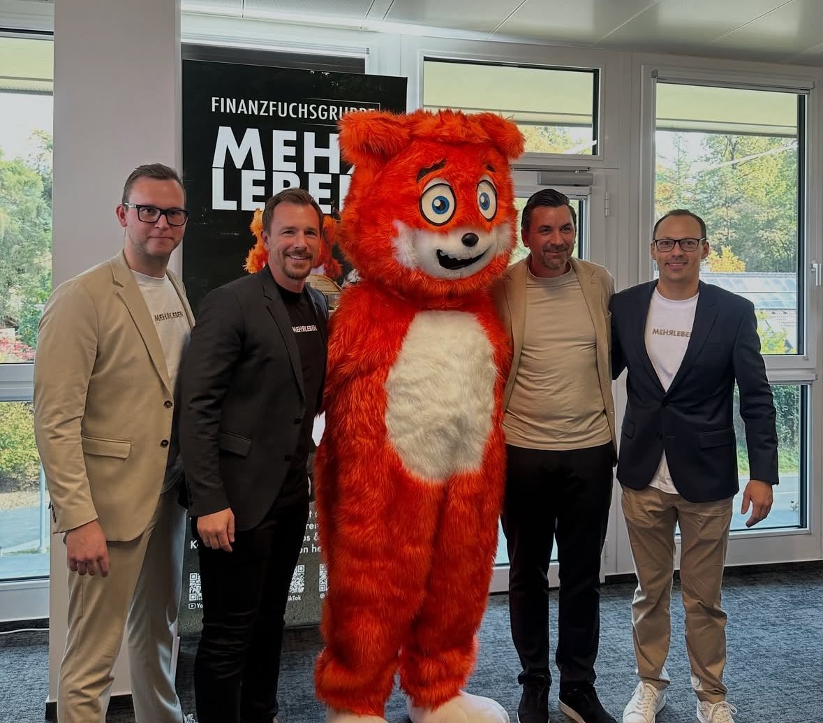 Vier Männer stehen beim „Elevator Pitch“ der Jungen Wirtschaft Steiermark neben einem großen orangefarbenen Maskottchen in einem Raum, im Hintergrund ist ein Werbebanner zu sehen.