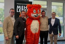 Vier Männer stehen beim „Elevator Pitch“ der Jungen Wirtschaft Steiermark neben einem großen orangefarbenen Maskottchen in einem Raum, im Hintergrund ist ein Werbebanner zu sehen.