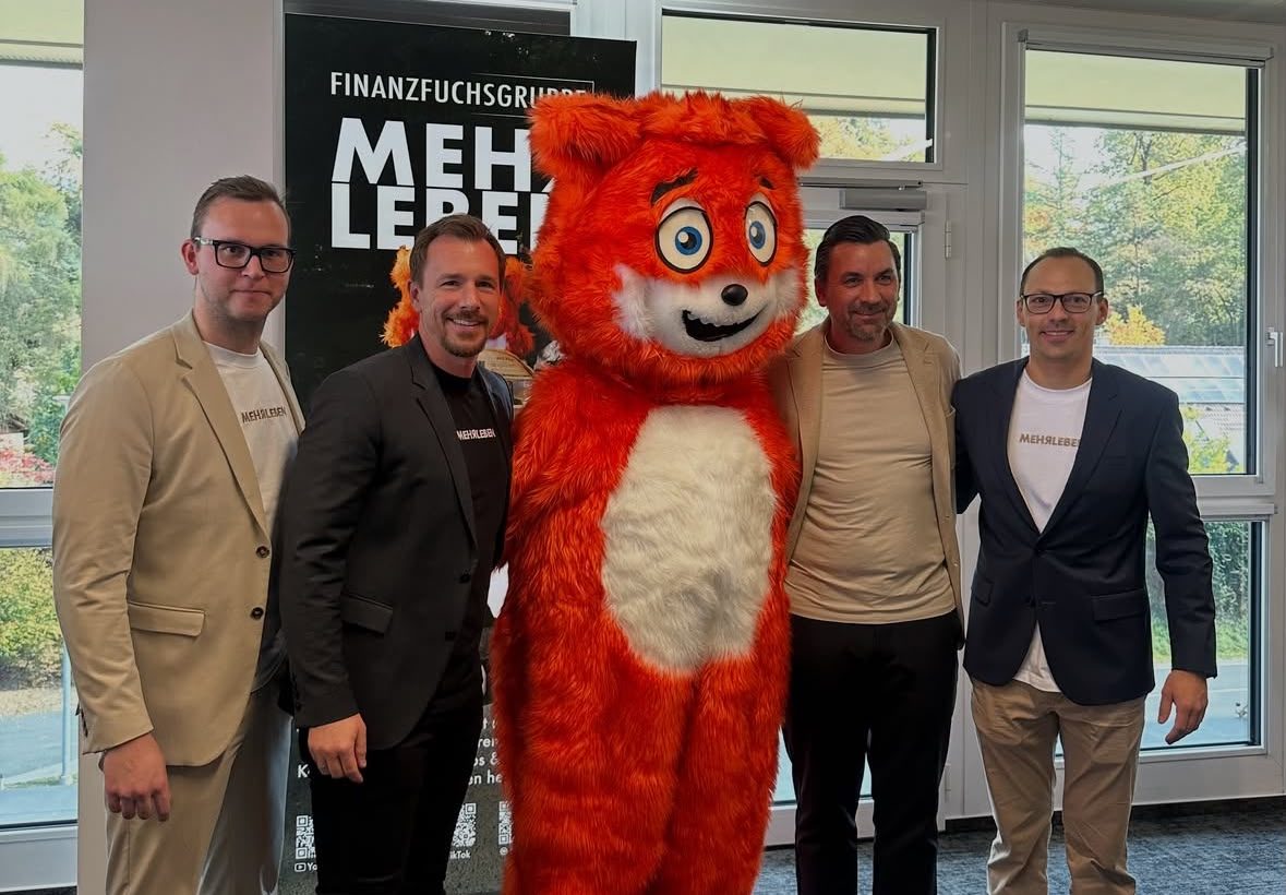 Vier Männer stehen beim „Elevator Pitch“ der Jungen Wirtschaft Steiermark neben einem großen orangefarbenen Maskottchen in einem Raum, im Hintergrund ist ein Werbebanner zu sehen.