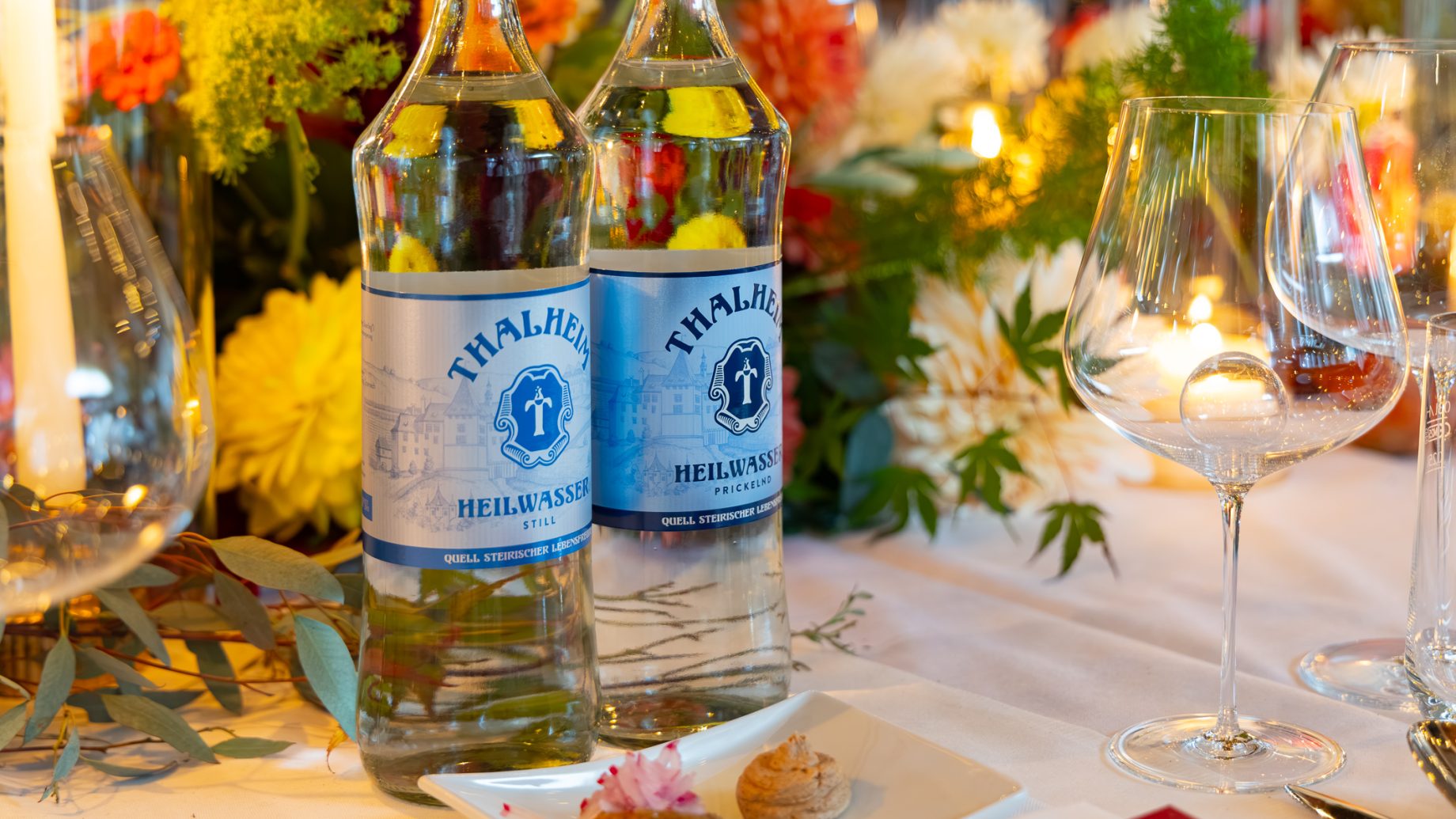 Zwei Glasflaschen Thalheim-Mineralwasser auf einem Tisch, der für „Der gedeckte Ort, Band 3“ gedeckt ist, mit Blumenarrangements, Weingläsern und Häppchen.