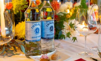 Zwei Glasflaschen Thalheim-Mineralwasser auf einem Tisch, der für „Der gedeckte Ort, Band 3“ gedeckt ist, mit Blumenarrangements, Weingläsern und Häppchen.