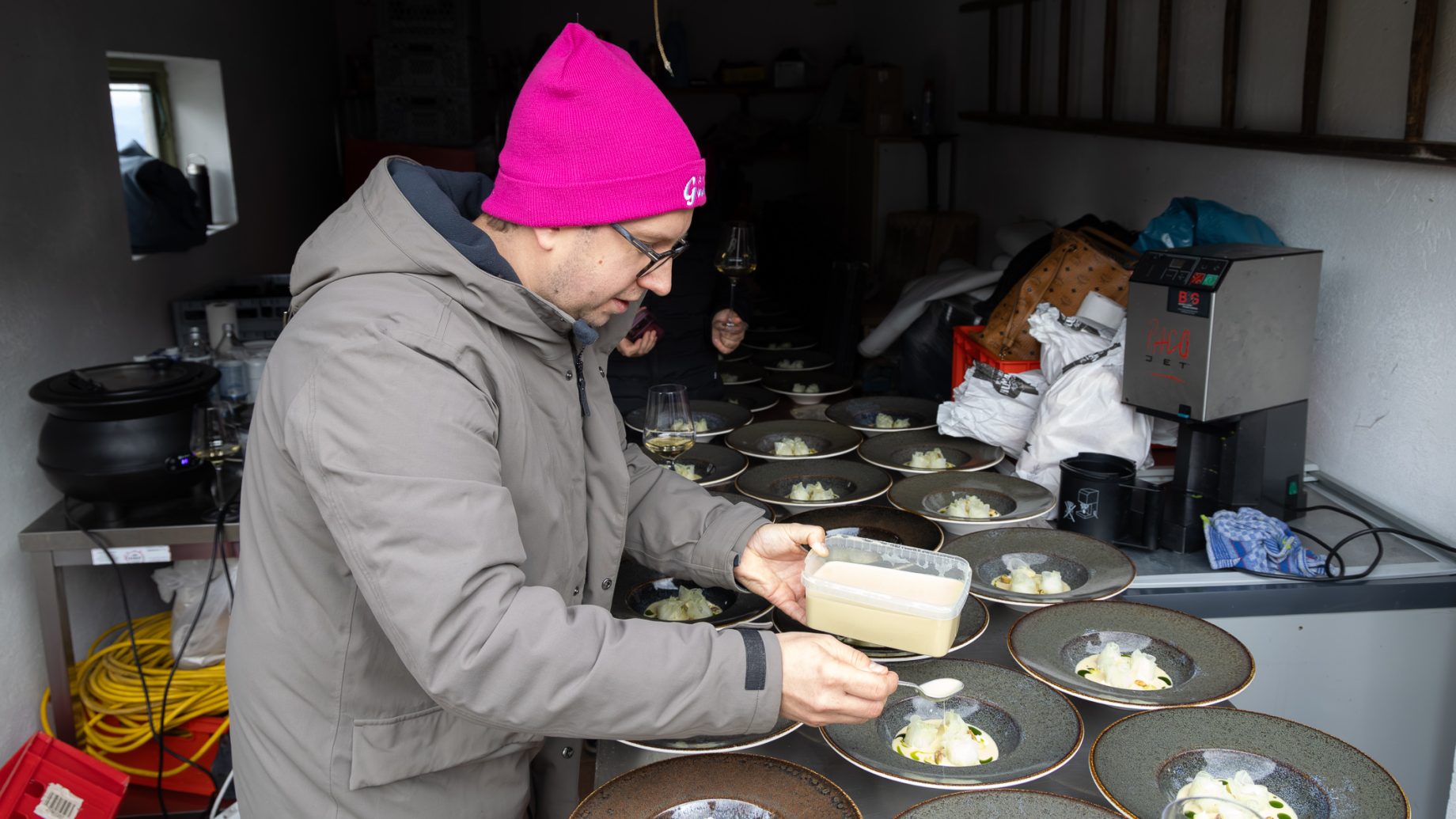 Eine Person mit einer rosa Mütze und einem grauen Mantel löffelt bei „Der gedeckte Ort Vol. 3“ Soße auf die angerichteten Speisen.