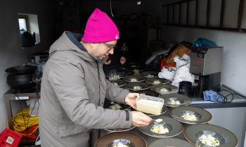 Eine Person mit einer rosa Mütze und einem grauen Mantel löffelt bei „Der gedeckte Ort Vol. 3“ Soße auf die angerichteten Speisen.