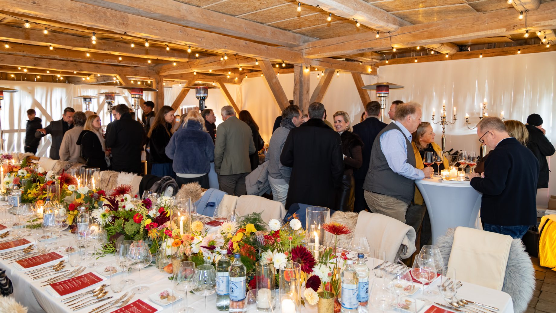 Die Gäste unterhalten sich auf einer Cocktailparty im Rahmen von „Der gedeckte Ort, Band 3“ in einem rustikalen Holzgebäude, in dem ein langer Esstisch mit Blumenarrangements geschmückt ist.