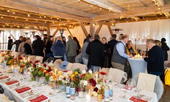 Die Gäste unterhalten sich auf einer Cocktailparty im Rahmen von „Der gedeckte Ort, Band 3“ in einem rustikalen Holzgebäude, in dem ein langer Esstisch mit Blumenarrangements geschmückt ist.