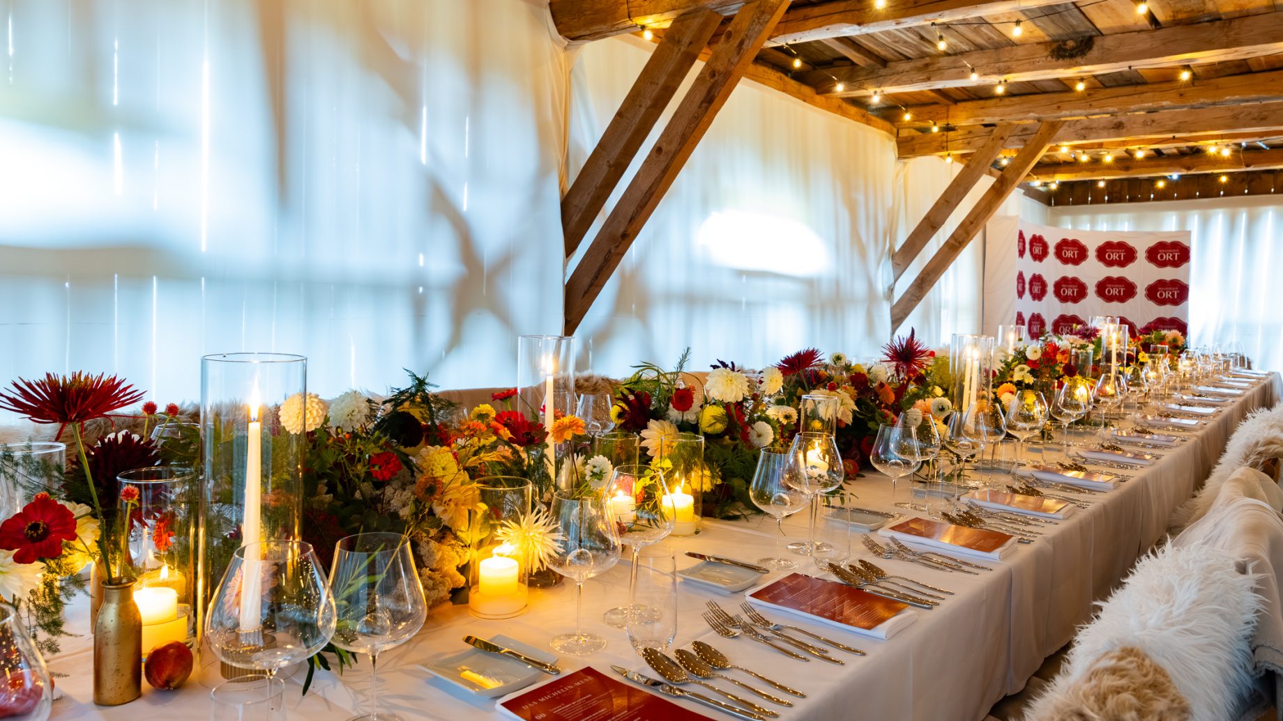 Ein langes Banketttisch-Set aus der Reihe „Der gedeckte Ort“, Band 3, gedeckt für ein festliches Abendessen mit Blumengestecken, Kerzen, Gläsern und roten Speisekarten.
