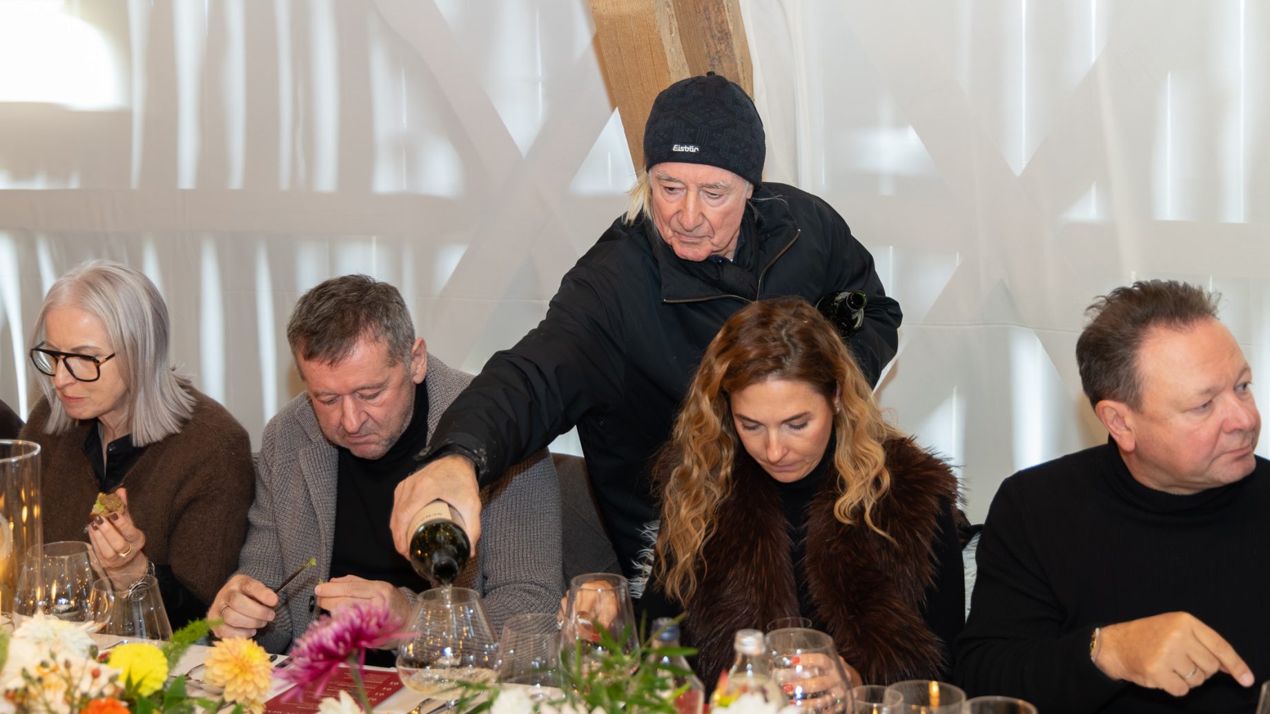 Ein Mann in schwarzer Jacke und Mütze schenkt einer Frau am Esstisch von „Der gedeckte Ort, Band 3“ Wein ein, umgeben von anderen Gästen.