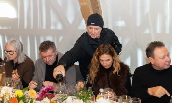 Ein Mann in schwarzer Jacke und Mütze schenkt einer Frau am Esstisch von „Der gedeckte Ort, Band 3“ Wein ein, umgeben von anderen Gästen.