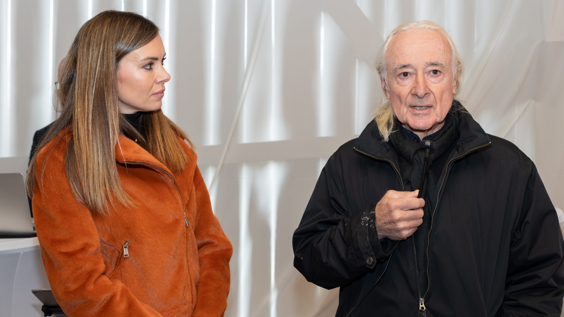 Bei „Der gedeckte Ort Vol. 3“ steht eine Frau in einer orangefarbenen Jacke neben einem Mann mit langen weißen Haaren, der ein Mikrofon in der Hand hält.