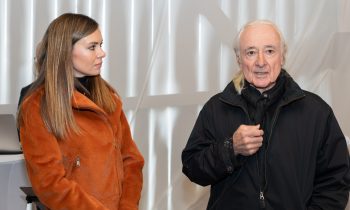 Bei „Der gedeckte Ort Vol. 3“ steht eine Frau in einer orangefarbenen Jacke neben einem Mann mit langen weißen Haaren, der ein Mikrofon in der Hand hält.