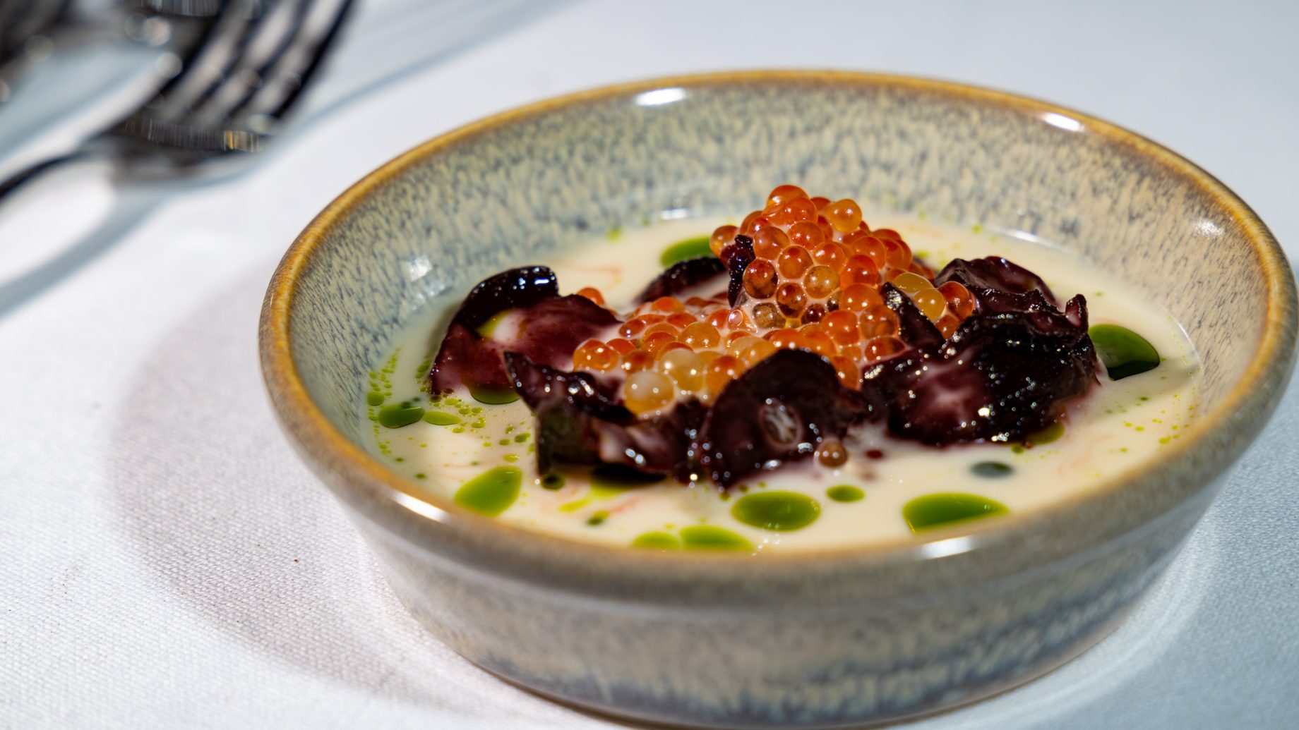 Eine kleine Schüssel cremige Suppe aus „Der gedeckte Ort“, Band 3, garniert mit Tintenfisch, Lachsrogen und einigen Tropfen grünem Kräuteröl.
