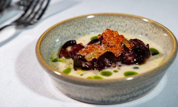 Eine kleine Schüssel cremige Suppe aus „Der gedeckte Ort“, Band 3, garniert mit Tintenfisch, Lachsrogen und einigen Tropfen grünem Kräuteröl.