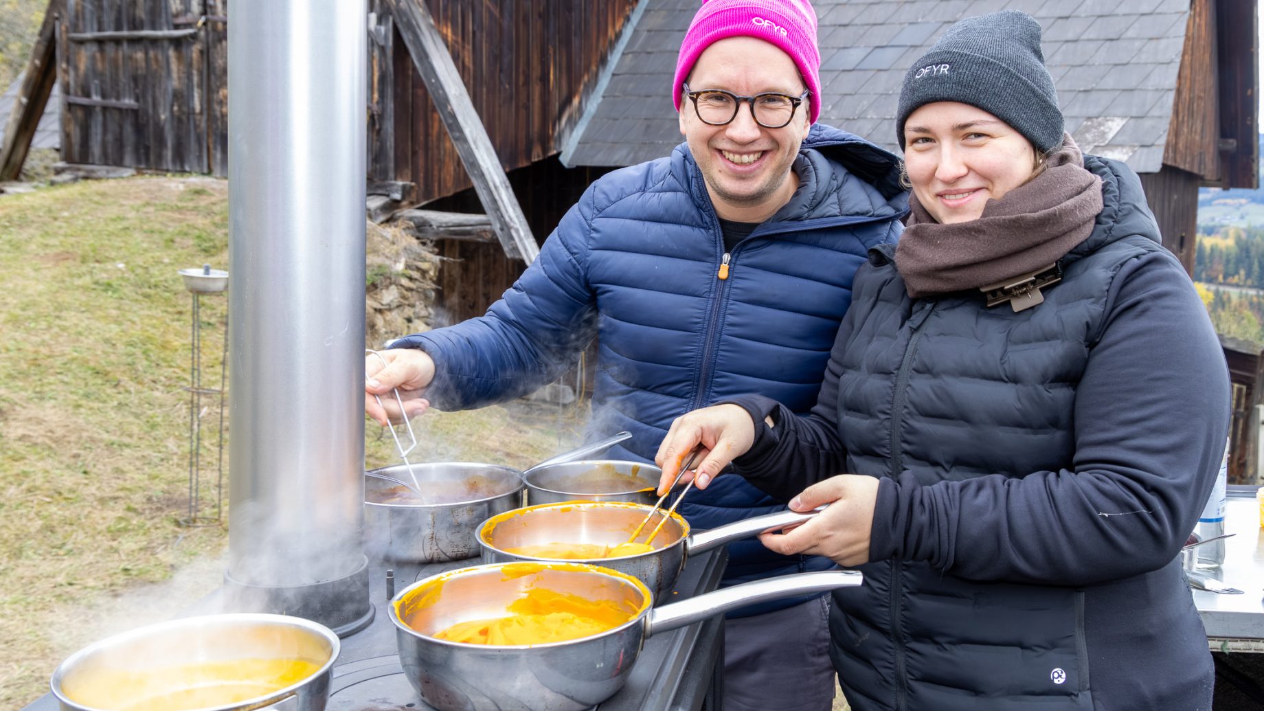 Ein Mann und eine Frau mit Wintermützen und Jacken kochen für „Der gedeckte Ort, Band 3“ in Töpfen auf einem Außenkocher.