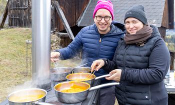 Ein Mann und eine Frau mit Wintermützen und Jacken kochen für „Der gedeckte Ort, Band 3“ in Töpfen auf einem Außenkocher.