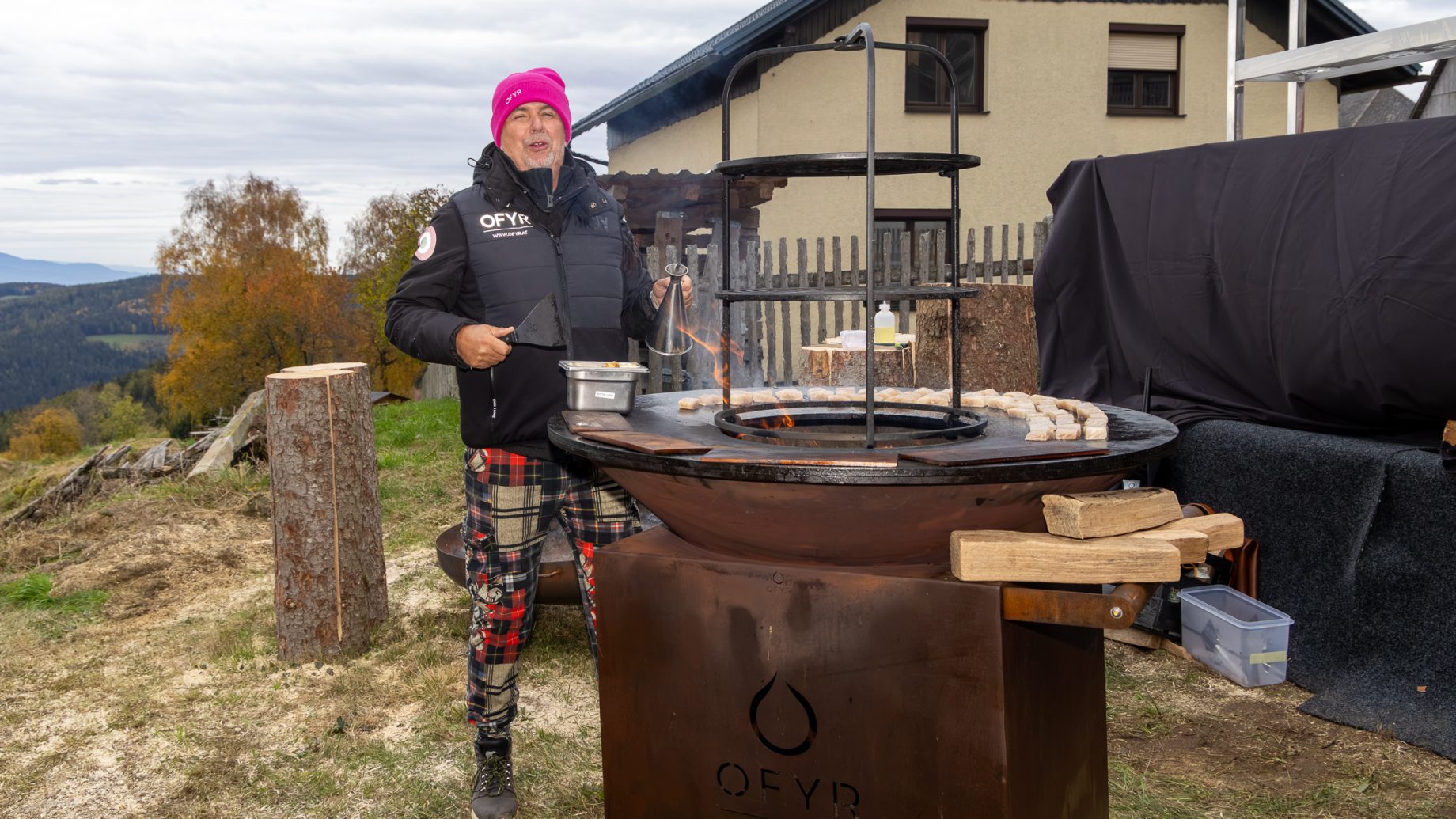 Ein Mann mit einer rosa Mütze steht neben einem runden OFYR-Holzkohlegrill und backt im Freien kleine runde Kuchen für „Der gedeckte Ort, Band 3“.