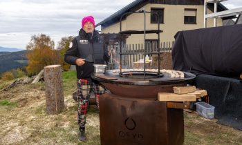 Ein Mann mit einer rosa Mütze steht neben einem runden OFYR-Holzkohlegrill und backt im Freien kleine runde Kuchen für „Der gedeckte Ort, Band 3“.