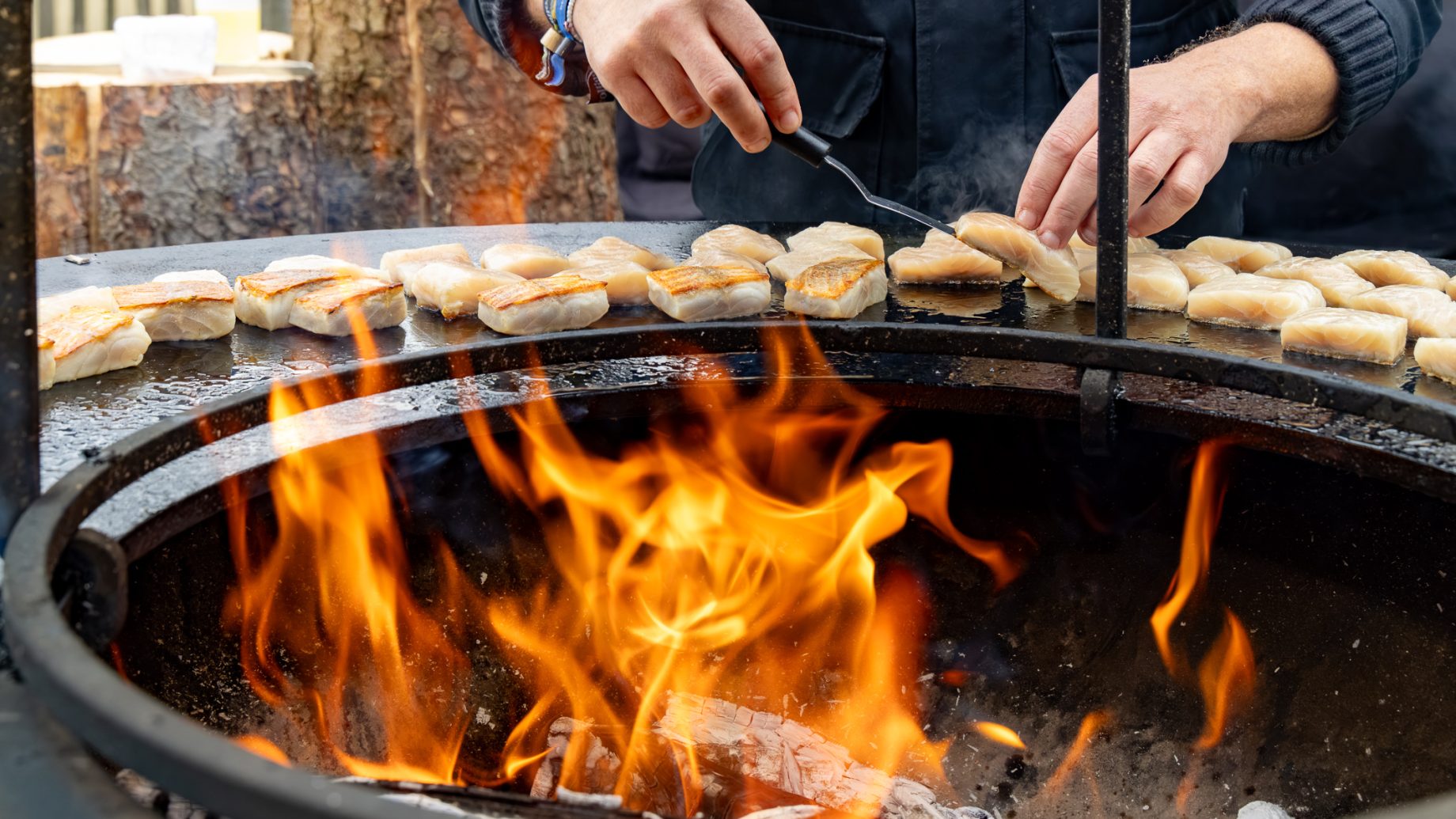 Ein Koch wendet mit einem Metallspatel Fischstücke, die auf einem runden Grill über offenem Feuer anbraten, wie in „Der gedeckte Ort“, Band 3, zu sehen ist.