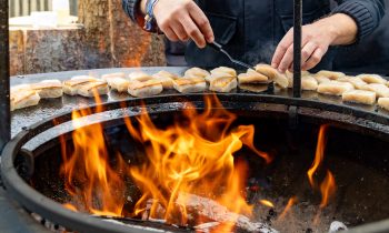 Ein Koch wendet mit einem Metallspatel Fischstücke, die auf einem runden Grill über offenem Feuer anbraten, wie in „Der gedeckte Ort“, Band 3, zu sehen ist.