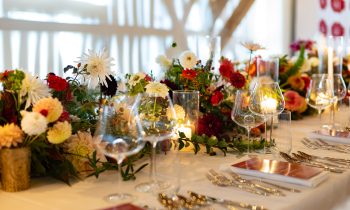 A Der gedeckte Ort, Band 3: Ein Festtagstisch mit Blumengestecken, brennenden Kerzen, Gläsern und Gedecken.