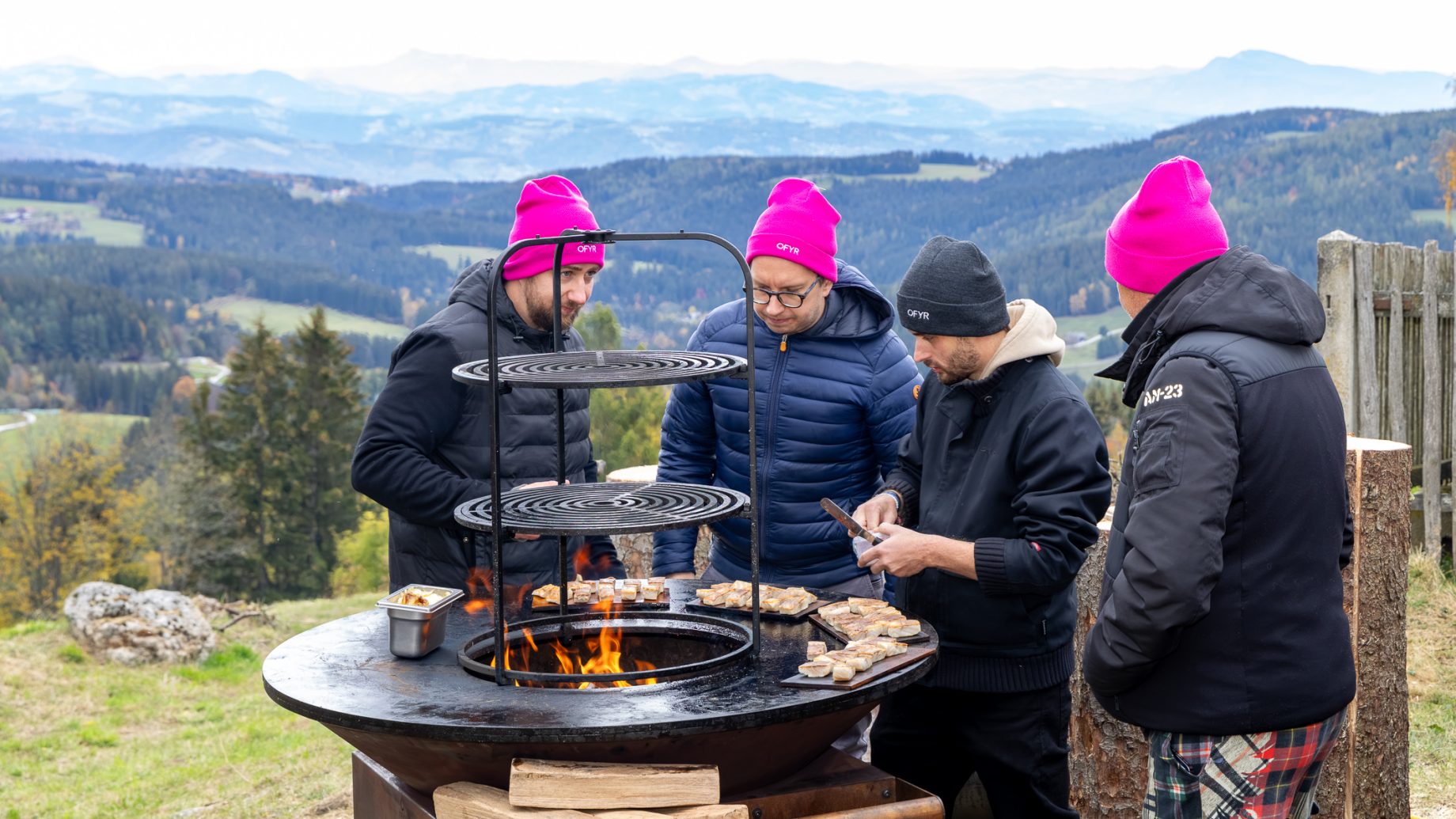 Vier Menschen mit Wintermützen braten Fleisch auf einer Feuerstelle im Freien bei „Der gedeckte Ort Vol. 3“ und blicken dabei auf eine Berglandschaft.