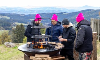 Vier Menschen mit Wintermützen braten Fleisch auf einer Feuerstelle im Freien bei „Der gedeckte Ort Vol. 3“ und blicken dabei auf eine Berglandschaft.