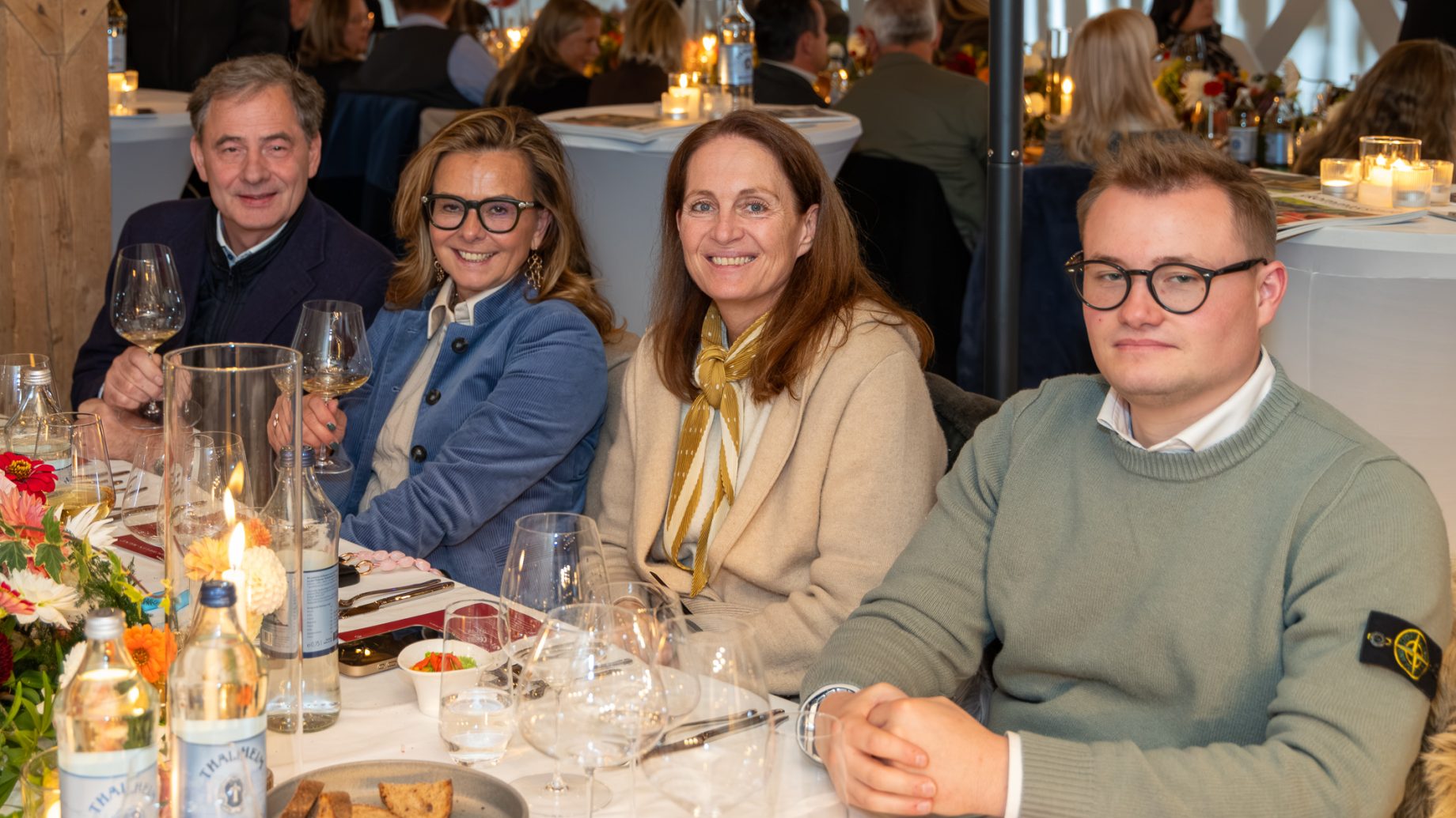 In „Der gedeckte Ort, Band 3“ sitzen vier Personen an einem Tisch in einem Restaurant, auf dem Weingläser, Wasserflaschen und Speisen stehen.
