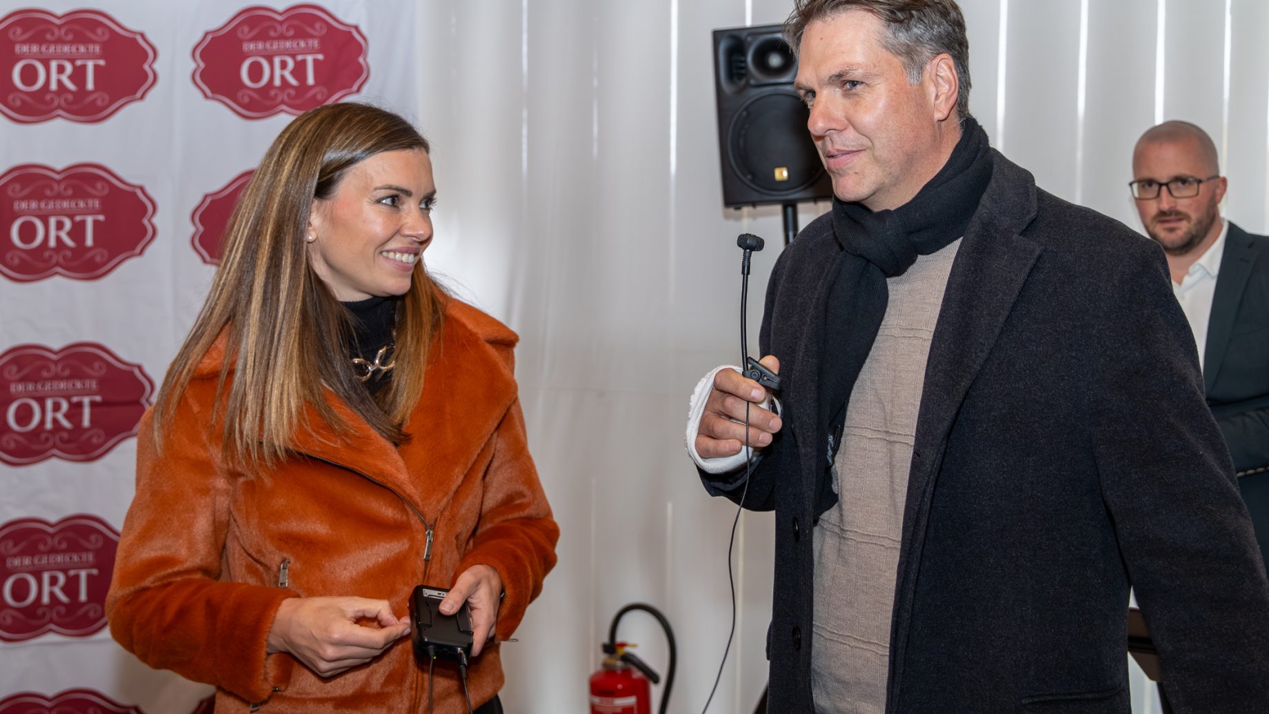 Eine Frau in einer orangefarbenen Jacke interviewt für „Der gedeckte Ort, Band 3“ einen Mann in einem schwarzen Mantel, der ein Mikrofon in der Hand hält; im Hintergrund ist ein weiterer Mann zu sehen.