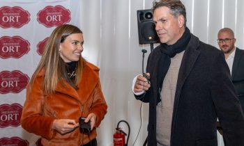 Eine Frau in einer orangefarbenen Jacke interviewt für „Der gedeckte Ort, Band 3“ einen Mann in einem schwarzen Mantel, der ein Mikrofon in der Hand hält; im Hintergrund ist ein weiterer Mann zu sehen.