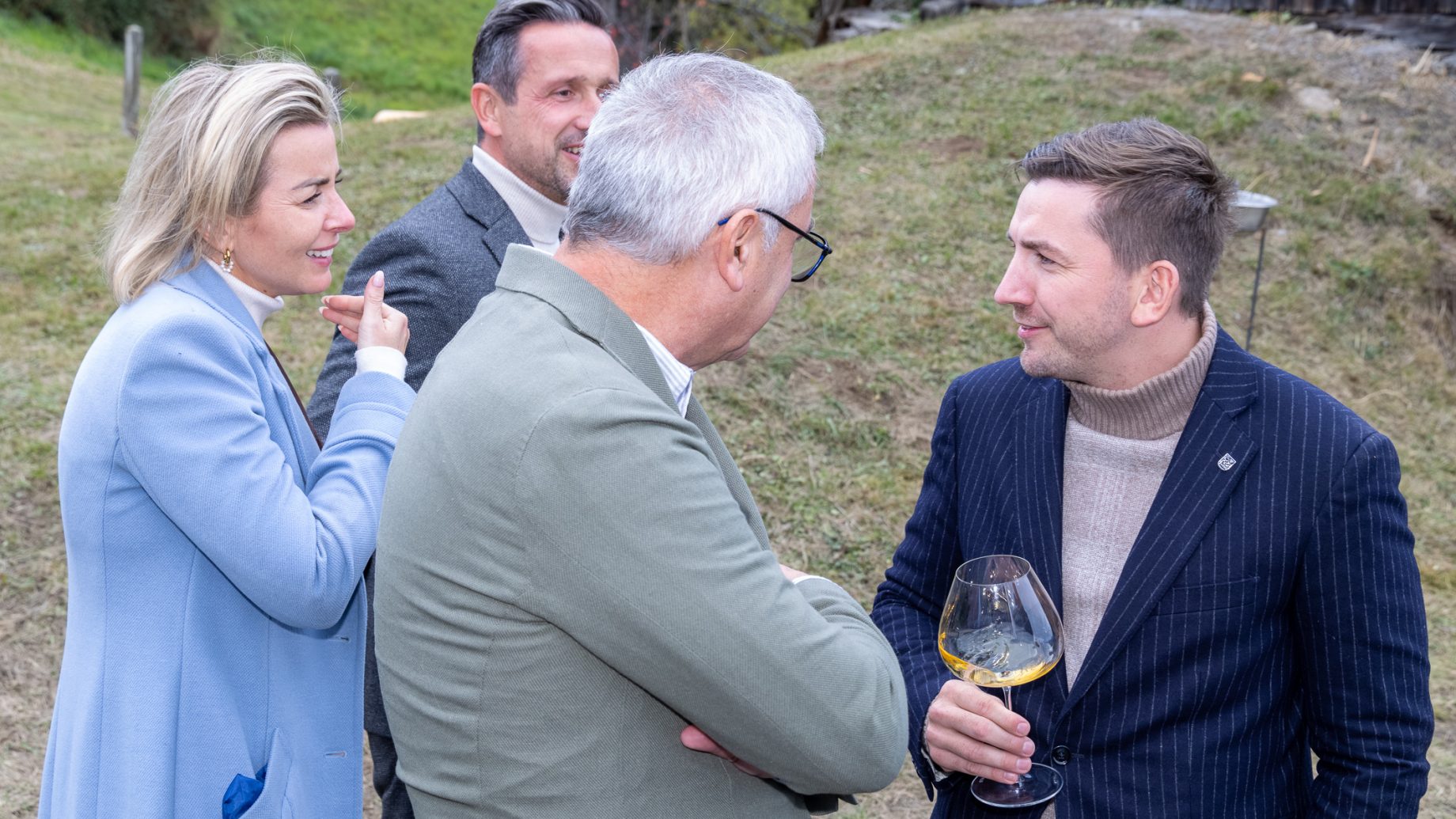 Vier Personen in Business-Casual-Kleidung genießen im Freien bei „Der gedeckte Ort Vol. 3“ Wein und gute Gespräche.