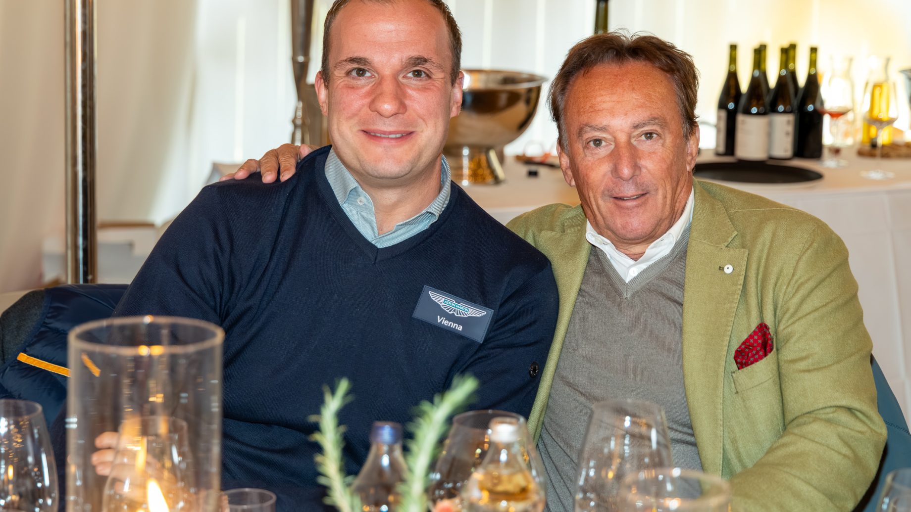 Zwei Männer sitzen gemeinsam an einem Tisch von „Der gedeckte Ort, Band 3“; der eine trägt einen marineblauen „Aston Martin Vienna“-Pullover, der andere einen grünen Blazer.