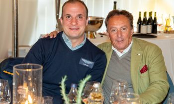 Zwei Männer sitzen gemeinsam an einem Tisch von „Der gedeckte Ort, Band 3“; der eine trägt einen marineblauen „Aston Martin Vienna“-Pullover, der andere einen grünen Blazer.