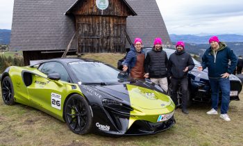 Vier Männer mit rosa Mützen stehen bei „Der gedeckte Ort Vol. 3“ neben einem grün-schwarzen McLaren, der an einem Hang vor einer Holzscheune geparkt ist.
