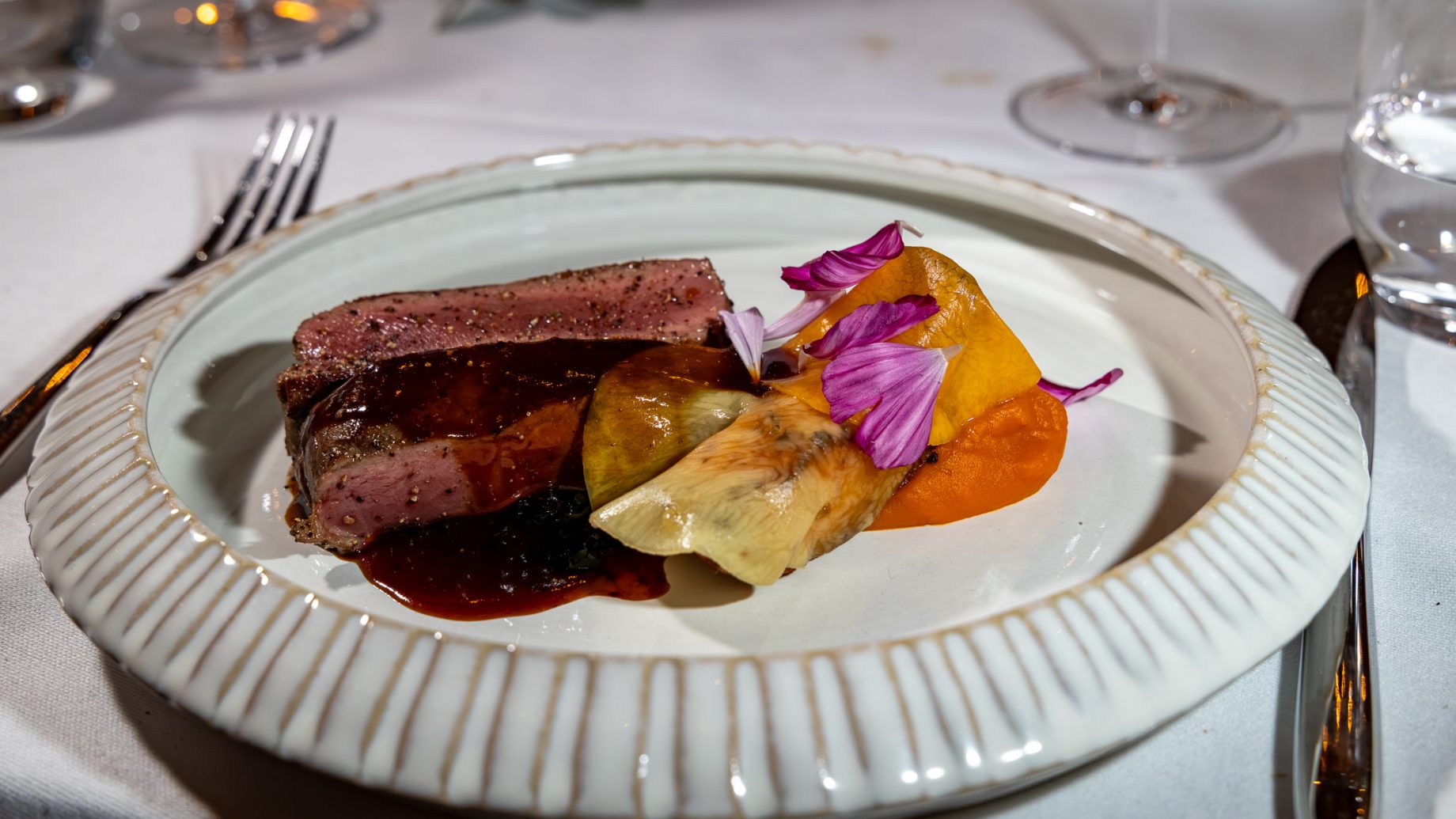 In Scheiben geschnittenes Steak mit Soße, Artischockenherzen und Gemüsepüree, garniert mit violetten Blütenblättern, serviert auf einem geriffelten Keramikteller von „Der gedeckte Ort, Band 3“.