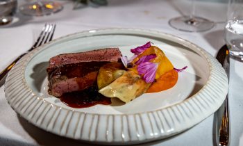 In Scheiben geschnittenes Steak mit Soße, Artischockenherzen und Gemüsepüree, garniert mit violetten Blütenblättern, serviert auf einem geriffelten Keramikteller von „Der gedeckte Ort, Band 3“.