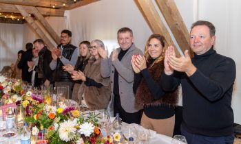 Eine Gruppe von Menschen an einem langen Esstisch für „Der gedeckte Ort, Band 3“, umgeben von Blumenarrangements und Glaswaren, die alle in die Hände klatschen.